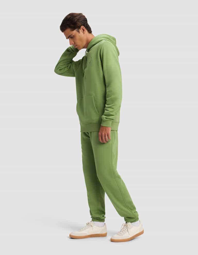 A man wears the Cozy Earth Men's Cityscape 2.0 Hoodie & Sweatpant Set in light green, paired with white sneakers with tan soles, standing against a plain light background and touching the back of his neck.|Color:Clover