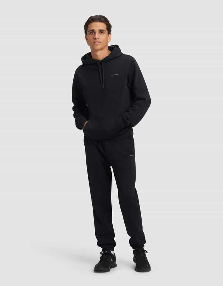 A young man faces forward in the Cozy Earth Men's Cityscape 2.0 Hoodie & Sweatpant Set, paired with black sneakers. His hands are in the hoodie pocket as he stands against a plain light gray background.|Color:Jet Black