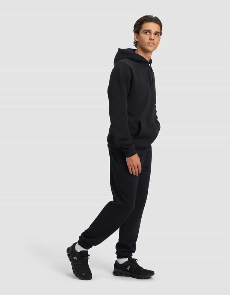 A young man models the Cozy Earth Men's Cityscape 2.0 Hoodie & Sweatpant Set in black, standing against a plain light gray background with one hand in his pocket, looking slightly to the side.|Color:Jet Black