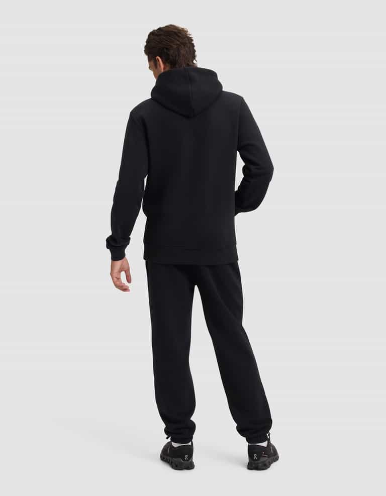 A person faces away from the camera, wearing Cozy Earth's Men's Cityscape 2.0 Hoodie & Sweatpant Set in black, with matching sneakers against a plain light gray background.|Color:Jet Black