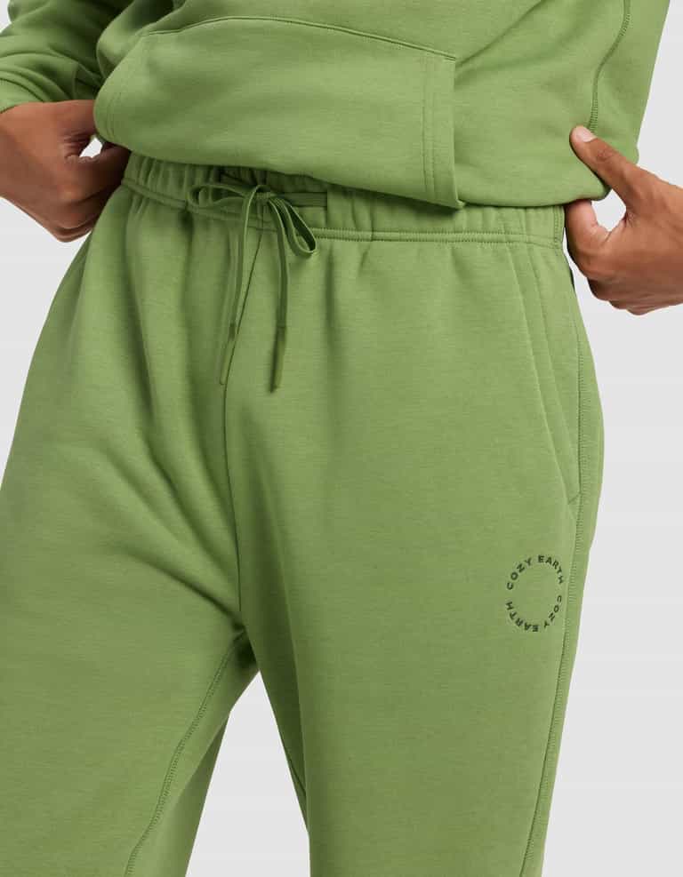 A person wears Cozy Earth Men's Cityscape 2.0 Sweatpant in light green with an elastic drawstring waistband, a small circular logo by the left pocket, and a matching sweatshirt, one hand in the pocket.|Color:Clover