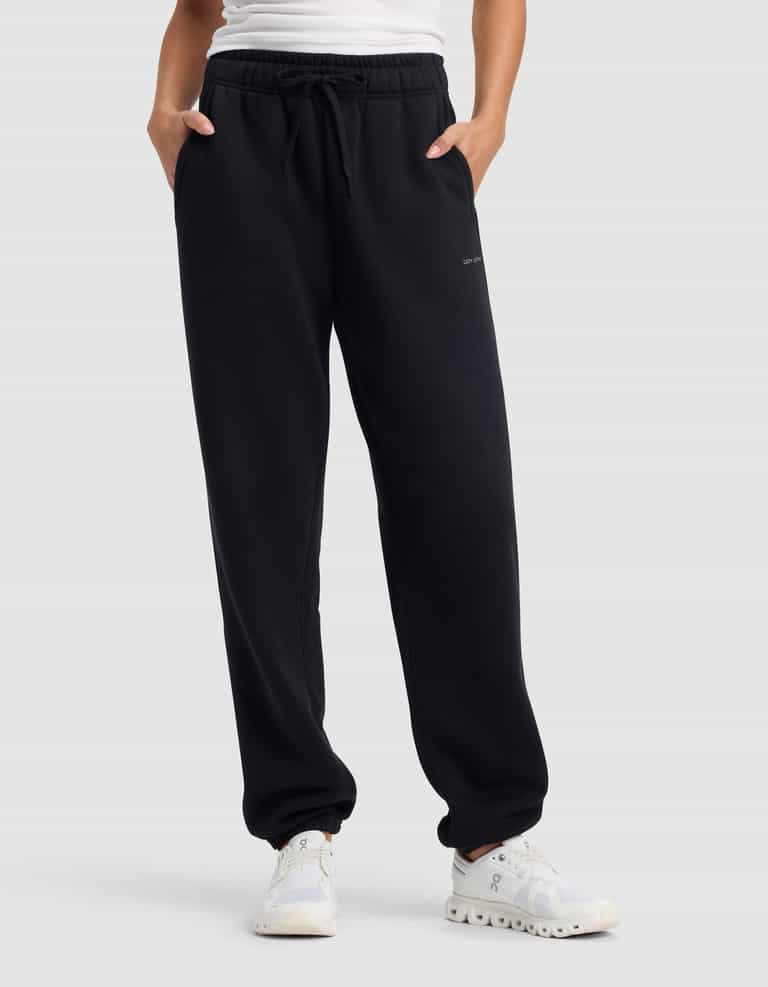 A person wearing Cozy Earth's Women’s Cityscape 2.0 Sweatpant in black, paired with a white shirt and white sneakers, stands with hands in pockets against a plain gray background.|Color:Jet Black