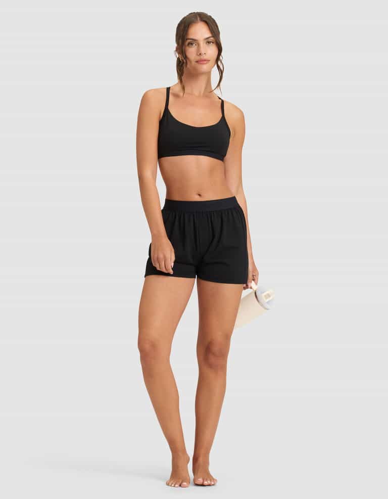 A woman stands barefoot against a plain background, wearing Cozy Earth's Women's First Layer Pajama Boxer Short in black and a matching black sports bra. She holds a rolled-up yoga mat in her right hand, looking forward with a relaxed expression.|Color:Jet Black