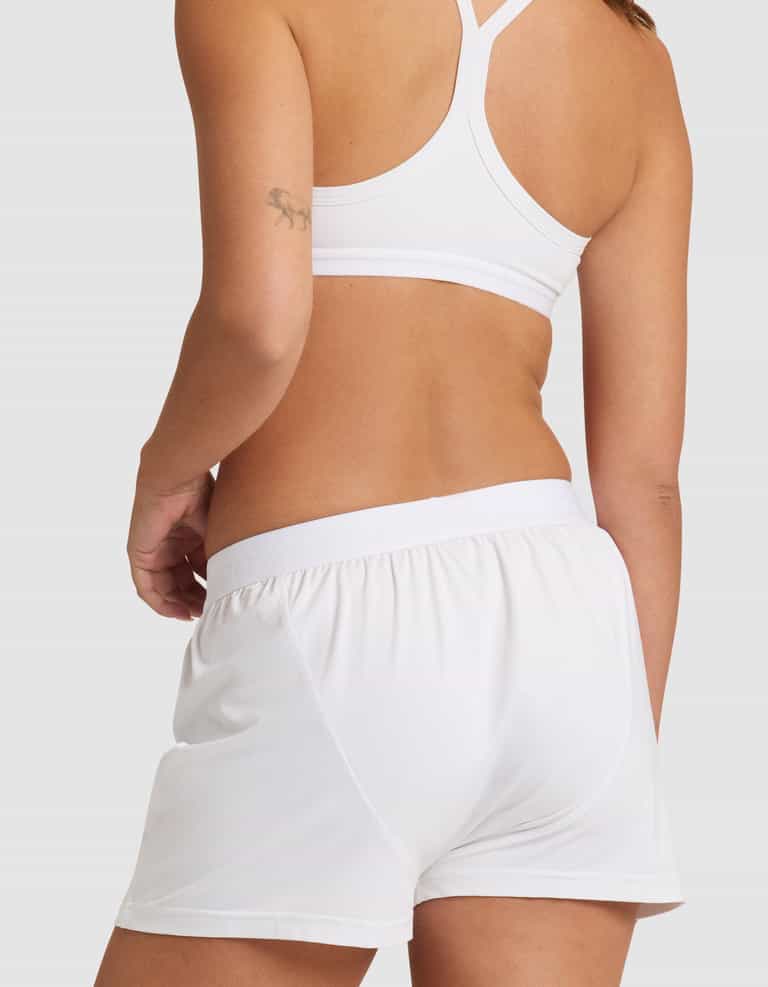 A woman is shown from the back against a plain light background, wearing Cozy Earth's Women's First Layer Pajama Boxer Short in white and a matching sports bra, with a small tattoo visible on her upper arm.|Color:White