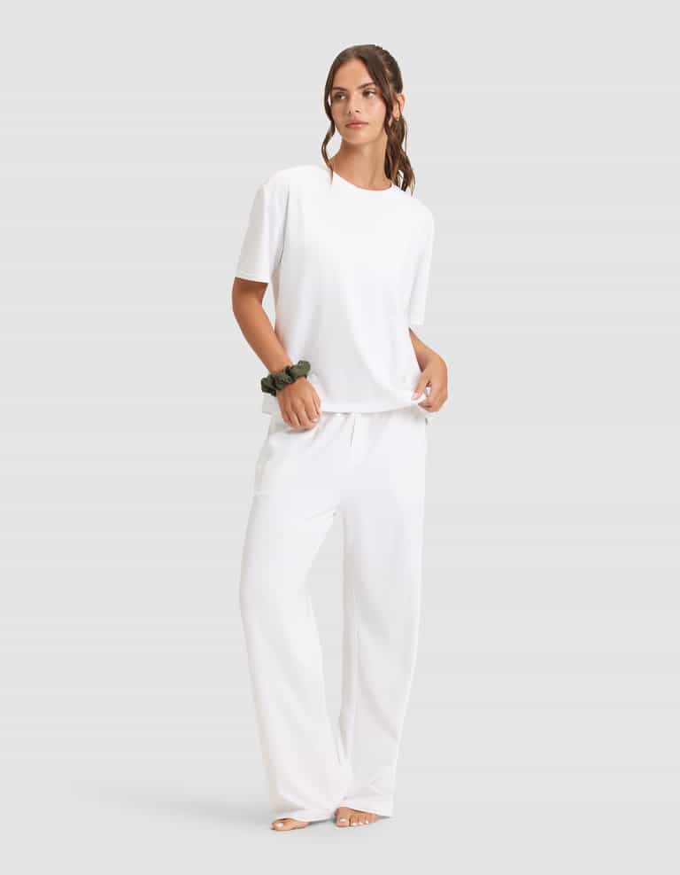 A woman with long brown hair tied back stands barefoot on a light background, wearing the Cozy Earth Women's First Layer Pajama Short Sleeve Tee & Pant Set in white, with a green scrunchie on her wrist.|Color:White