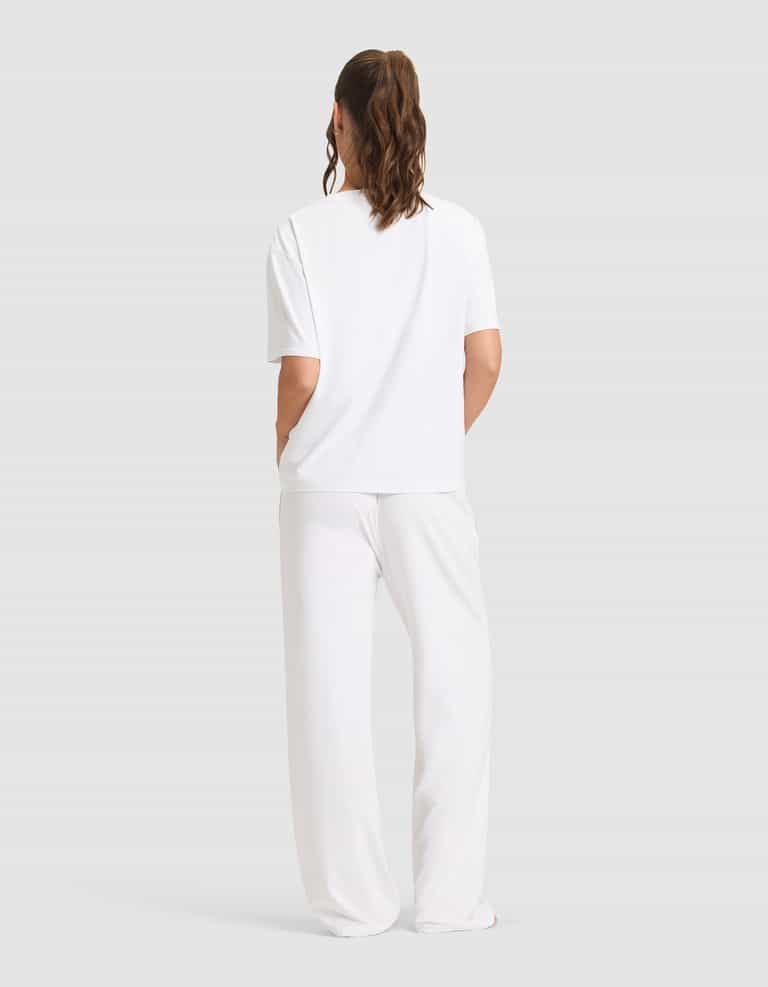 A person stands facing away, wearing the Cozy Earth Women's First Layer Pajama Short Sleeve Tee & Pant Set in white against a plain light gray background.|Color:White