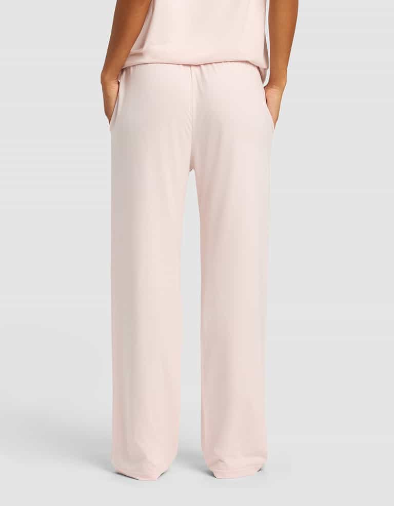 A woman wearing Cozy Earth Women's First Layer Pajama Pants in light pink, hands in pockets, is shown from the back against a plain light gray background.|Color:Ballet