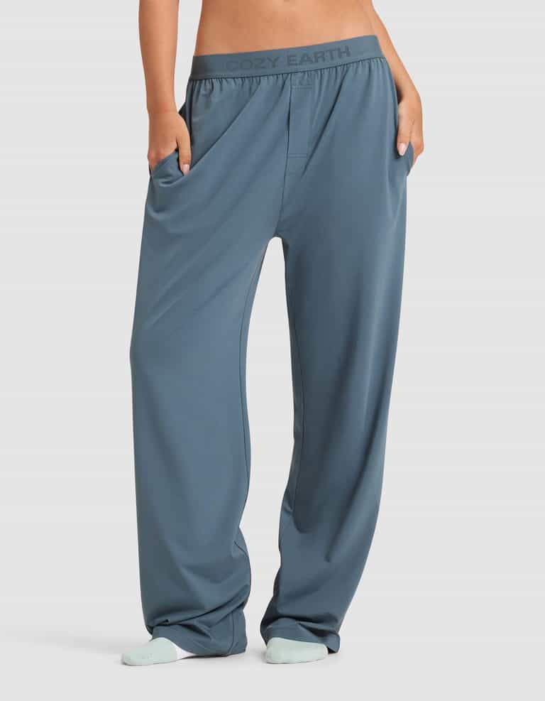 A person wearing Cozy Earth Women's First Layer Pajama Pant—loose-fitting, blue, with pockets and a branded waistband—stands with hands in the pockets and light blue socks.|Color:Dawn