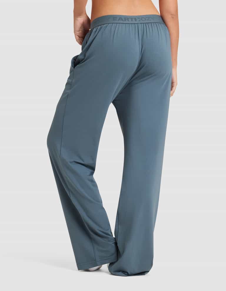A woman stands barefoot against a plain white background, shown from the back wearing Cozy Earth Women's First Layer Pajama Pant in a loose-fitting, high-waisted blue-gray style.|Color:Dawn