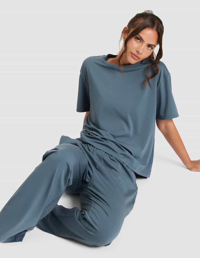 A woman with brown hair sits on the floor against a plain light background, wearing Cozy Earth's Women's First Layer Pajama Pant in blue with a matching loose short-sleeve top, looking at the camera with a relaxed expression.|Color:Dawn