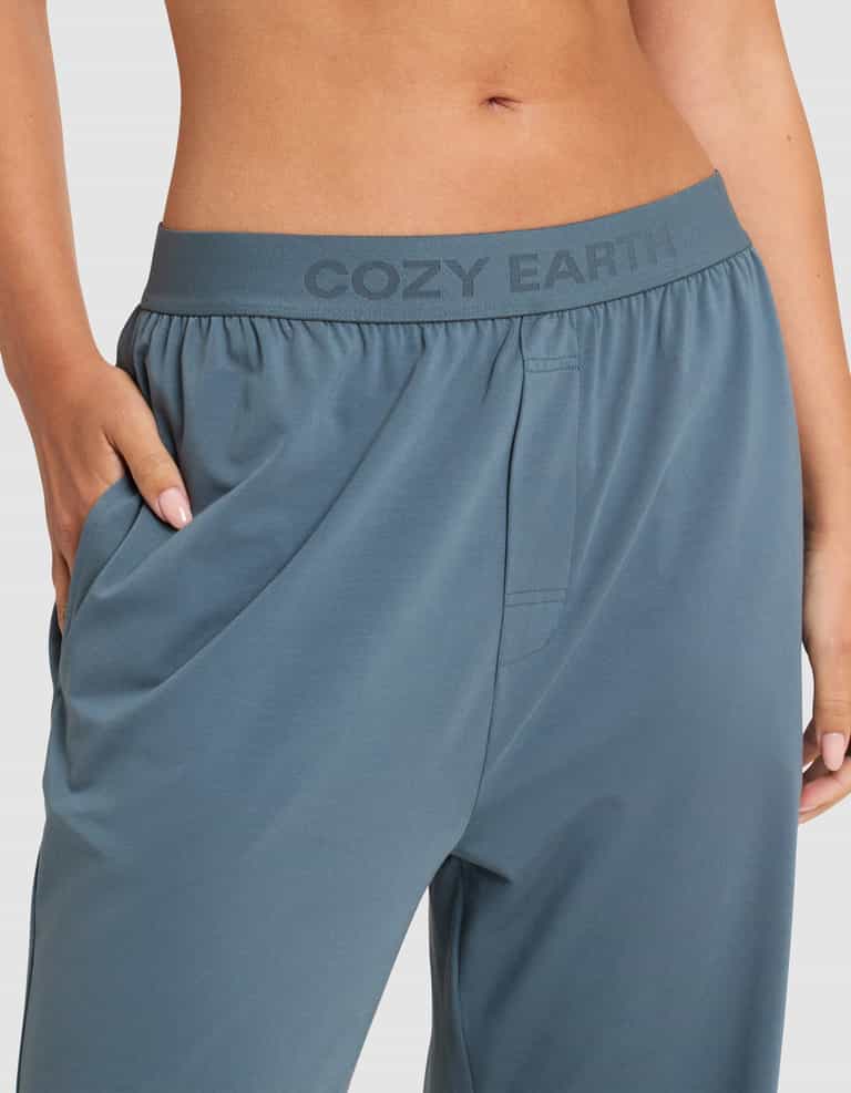 Close-up of a person wearing Cozy Earth Women's First Layer Pajama Pants with the brand name on the waistband. Their hand rests in the right pocket, and their midriff is visible against a plain background.|Color:Dawn