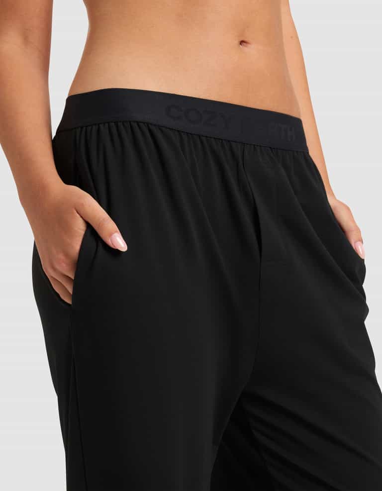 A woman models the Cozy Earth Women's First Layer Pajama Pant in black, hands in pockets, highlighting the branded waistband against a plain light gray background.|Color:Jet Black