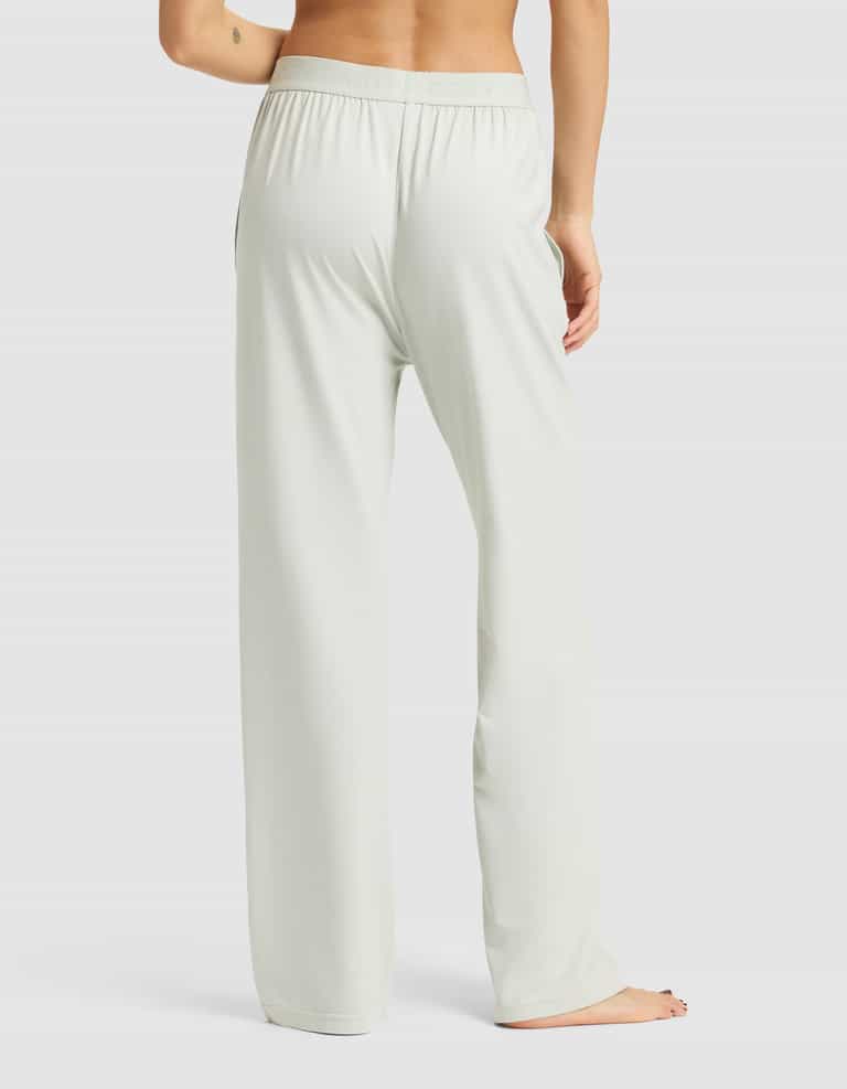 A woman stands barefoot on a white background, seen from the back, wearing Cozy Earth's Women's First Layer Pajama Pant in a light, loose fit with one arm slightly bent and visible.|Color:Laurel