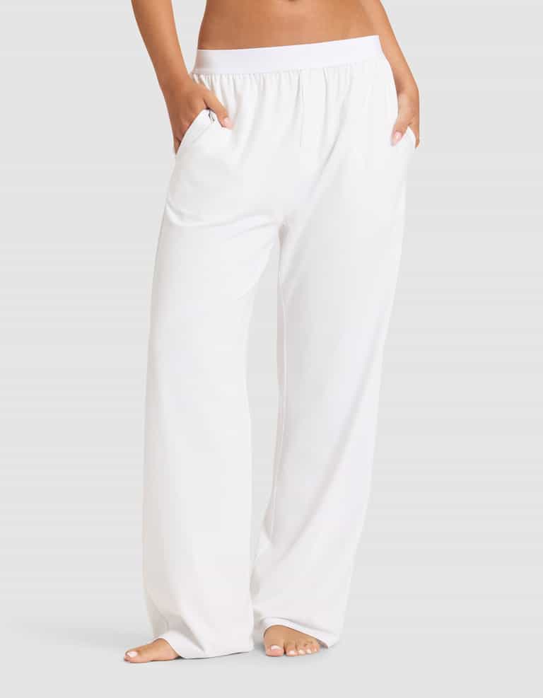 A woman stands barefoot, hands in pockets, wearing Cozy Earth's Women's First Layer Pajama Pant—loose-fitting white pants with an elastic waistband—against a plain light gray background.|Color:White