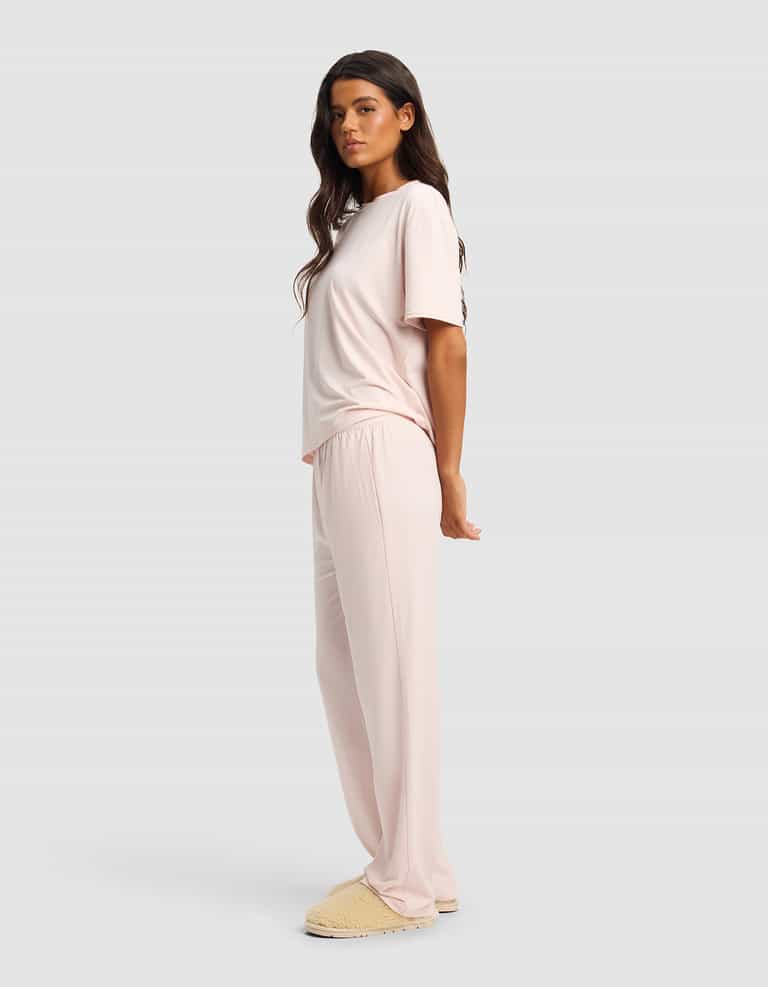 A woman stands sideways against a light gray background wearing the Cozy Earth Women's First Layer Pajama Short Sleeve Tee & Pant Set in light pink, paired with beige slippers.|Color:Ballet