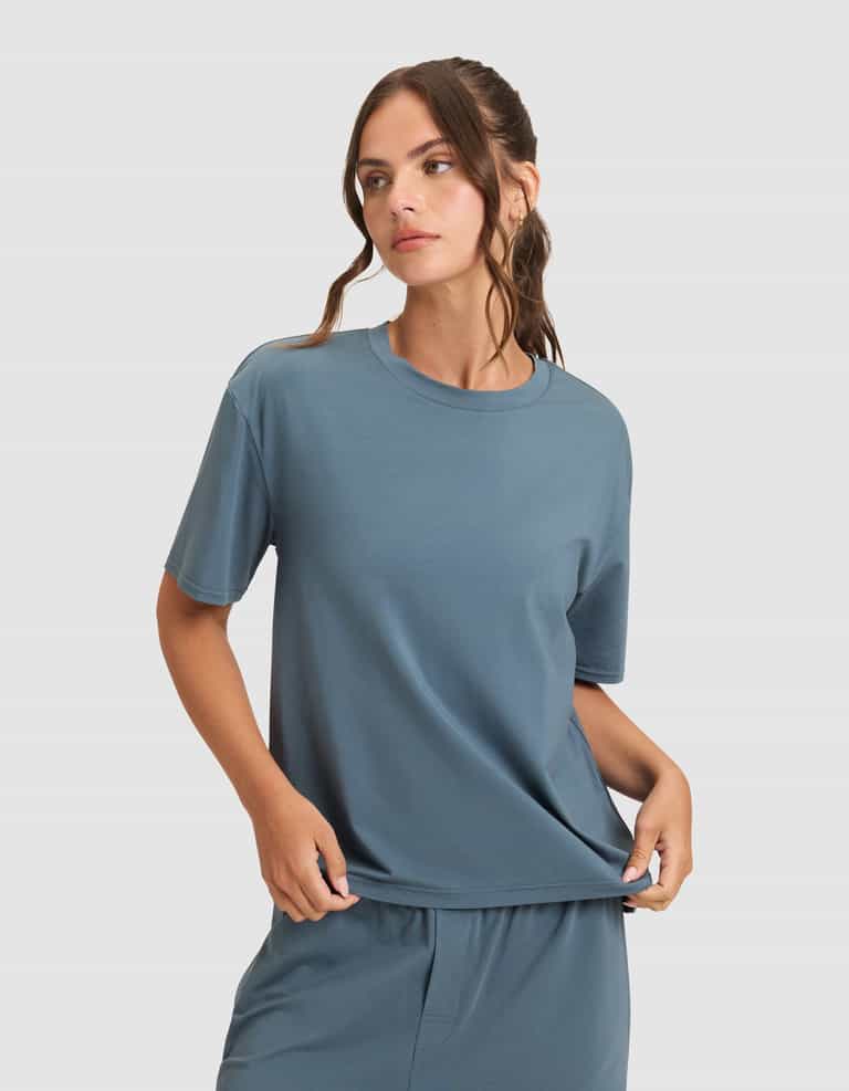 A woman wearing Cozy Earth's Women's First Layer Pajama Short Sleeve Tee and matching pants stands against a plain light gray background, looking to the side.|Color:Dawn