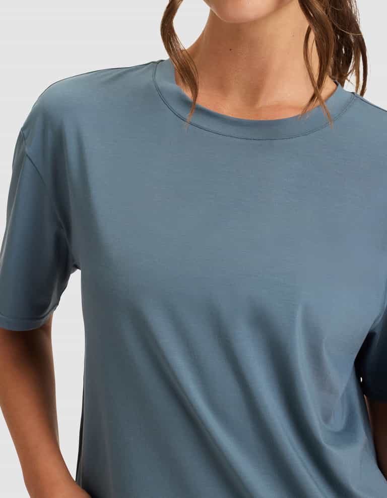 A woman stands with one hand on her hip, wearing the Cozy Earth Women's First Layer Pajama Short Sleeve Tee in light blue. Her head is partially visible, and her brown hair is partially tied back.|Color:Dawn