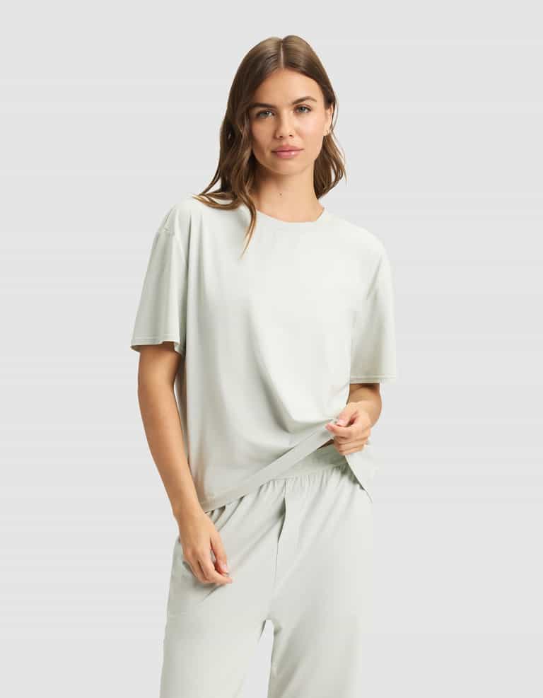 A woman with light brown hair wears the Cozy Earth Women's First Layer Pajama Short Sleeve Tee with matching pants, standing against a plain light gray background and smiling softly as she holds part of her shirt.|Color:Laurel