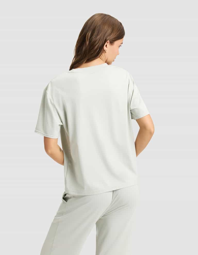 A woman with light brown hair is seen from behind wearing a Cozy Earth Women's First Layer Pajama Short Sleeve Tee in light grey, paired with matching pants, set against a plain light grey background.|Color:Laurel