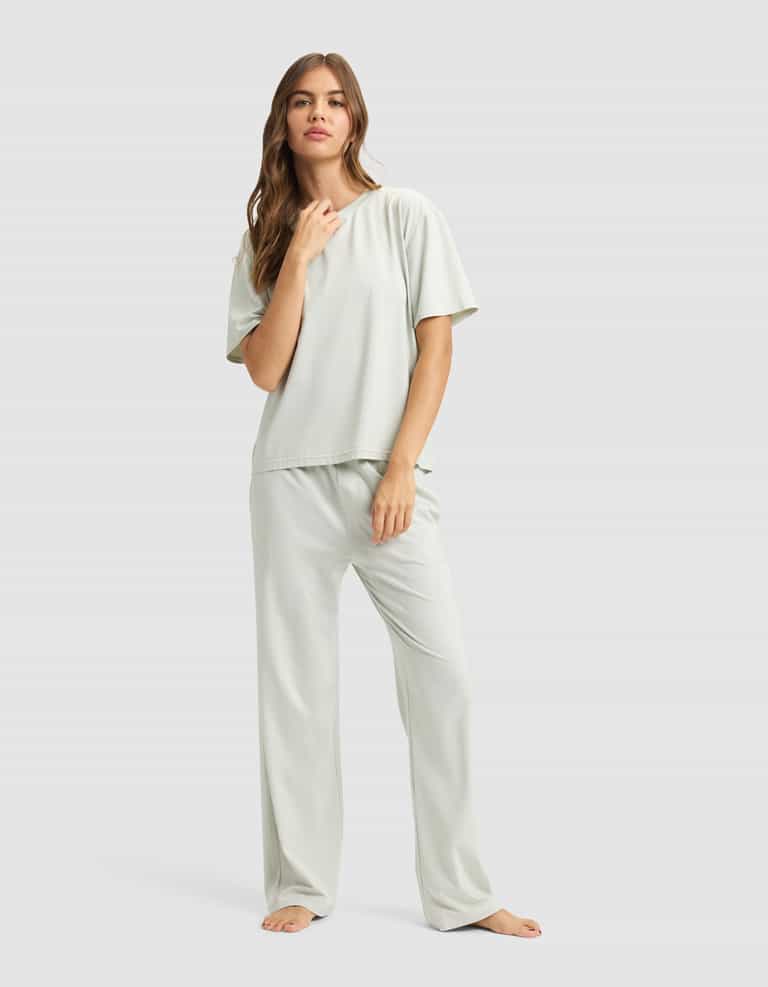 A woman with long, wavy hair stands barefoot against a plain background, confidently wearing Cozy Earth’s Women’s First Layer Pajama Short Sleeve Tee and matching pajama pants in a light color.|Color:Laurel