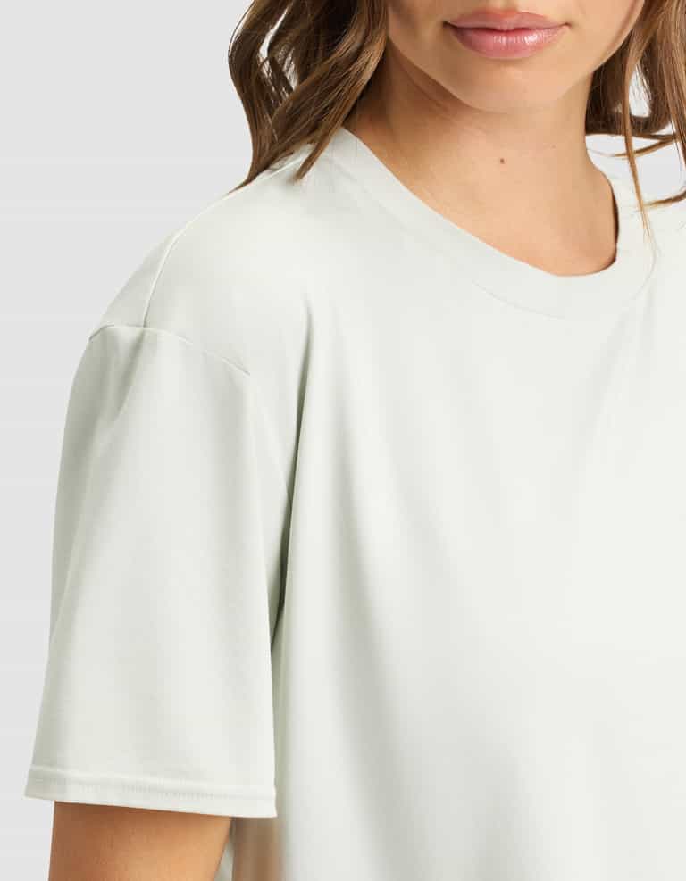 A woman wears the Cozy Earth Women's First Layer Pajama Short Sleeve Tee in a light color. Only her lower face, neck, shoulder, and upper torso are visible against a plain background.|Color:Laurel