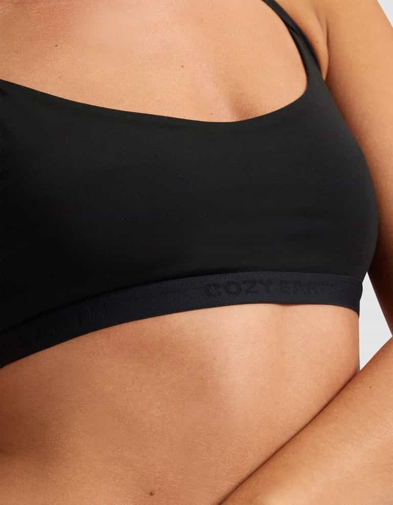 Close-up of a person wearing Cozy Earth's Women's First Layer Pajama Soft Bra from the Bra & Pant Set. The black bra has thin straps, a subtle embossed logo on the band, and is shown against a light plain background with arms crossed.|Color:Jet Black