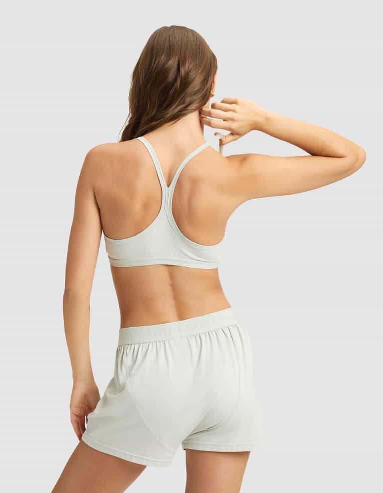 A woman with long brown hair faces away, wearing the Cozy Earth Women's First Layer PJ Soft Bra and matching shorts in light gray, standing against a plain light gray background with her right hand raised near her head.|Color:Laurel