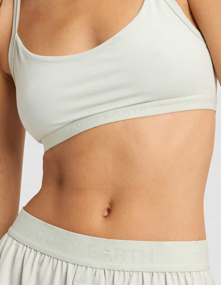 A person wears the Women's First Layer PJ Soft Bra by Cozy Earth, paired with matching shorts. The visible midsection and part of the chest and arm display "COZY EARTH" on the elastic bands against a plain background.|Color:Laurel