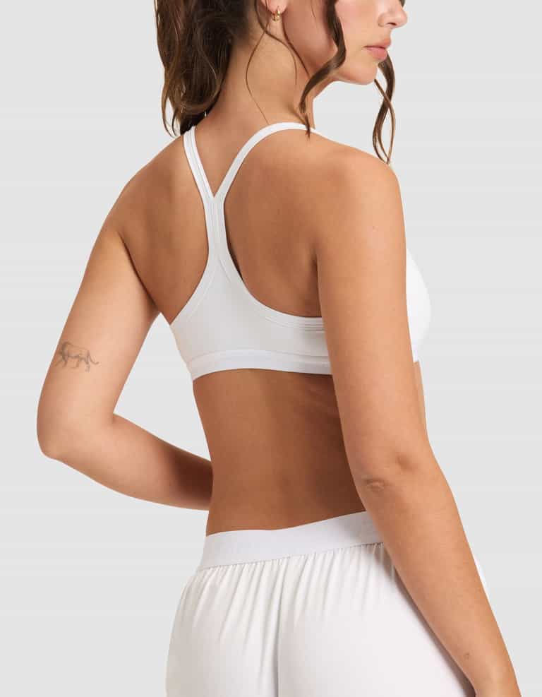 A woman with a ponytail and a small animal tattoo on her upper left arm wears the Cozy Earth Women's First Layer PJ Soft Bra in white, paired with white shorts, shown from the back against a plain light gray background.|Color:White