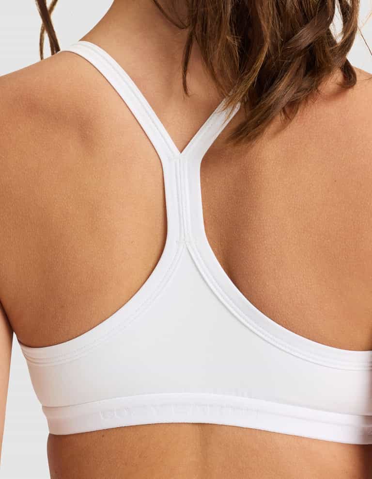 The image features the back view of a person wearing the Cozy Earth Women's First Layer PJ Soft Bra in white, showcasing its smooth racerback design. The person's hair is partially visible on the right side.|Color:White