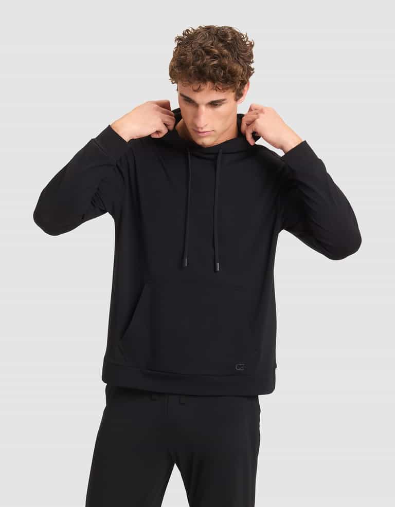 A young man with curly hair adjusts the hood of a black Cozy Earth Men's Brushed Bamboo Hoodie, paired with black pants, while standing against a plain light gray background. |Color:Black