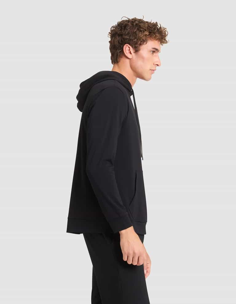 A young man with curly hair stands in profile, wearing Cozy Earth's Men's Brushed Bamboo Hoodie and black pants against a plain light gray background. |Color:Black