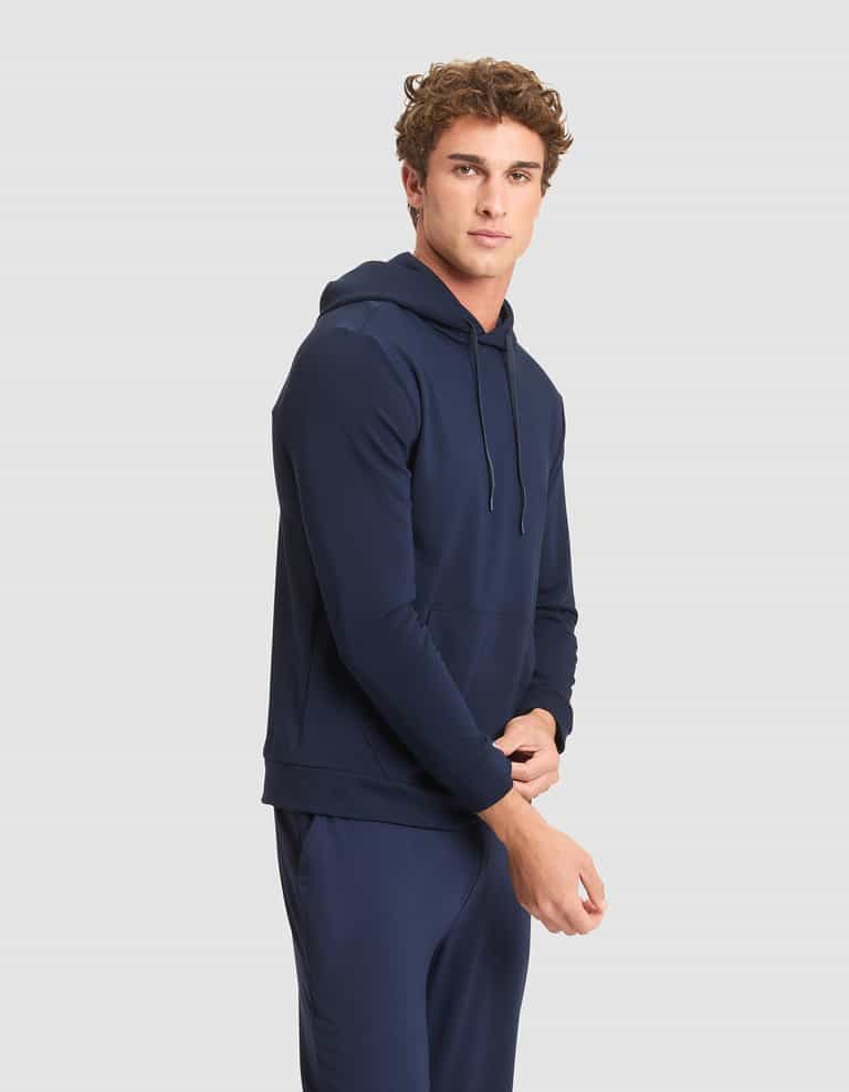 A young man with curly hair models the Cozy Earth Men's Brushed Bamboo Hoodie in navy blue, paired with matching pants, standing against a light gray background and looking at the camera with a neutral expression. |Color:Navy