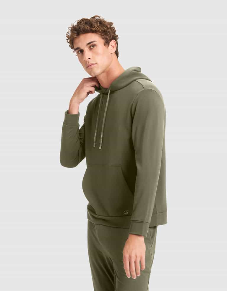 A young man with curly hair models the Cozy Earth Men's Brushed Bamboo Hoodie in olive green, paired with matching pants, standing against a plain light gray background and looking at the camera while touching his neck. |Color:Olive