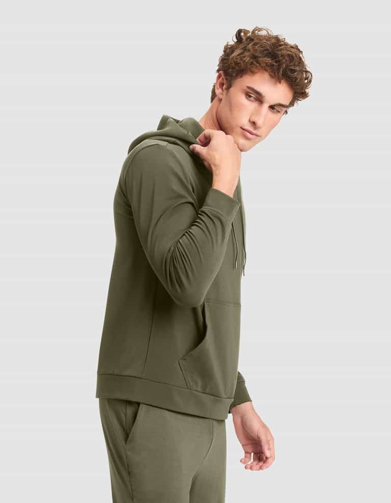 A young man with curly hair models the Cozy Earth Men's Brushed Bamboo Hoodie in olive green, paired with matching joggers, standing sideways and glancing over his shoulder against a plain light gray background. |Color:Olive