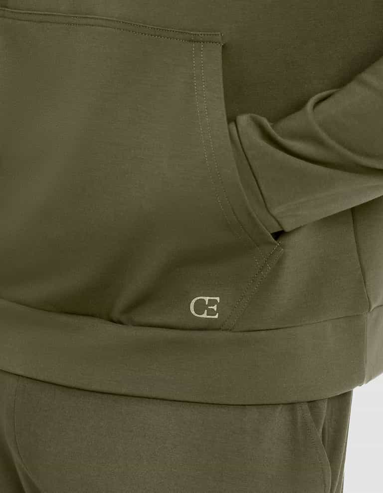 Close-up of a person wearing Cozy Earth's Men's Brushed Bamboo Hoodie in olive green, featuring matching pants and "CE" initials stitched in white near the front pocket. |Color:Olive
