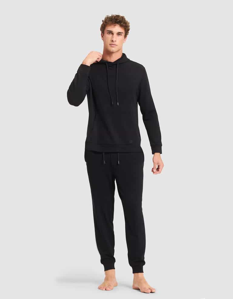 A person stands barefoot against a plain background, wearing the Cozy Earth Men's Brushed Bamboo Hoodie & Jogger Set in black, with their left hand near their neck and looking straight ahead with a neutral expression. |Color:Black