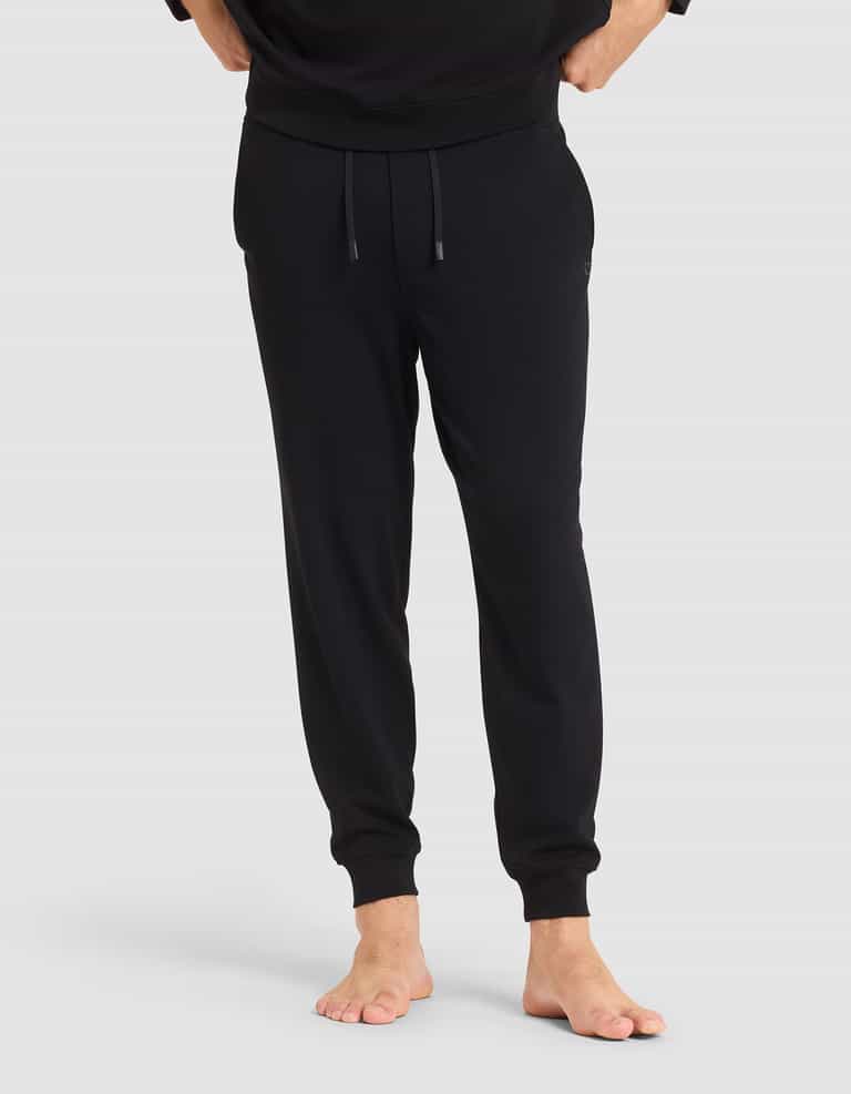 A person stands barefoot in Cozy Earth's Men's Brushed Bamboo Jogger Pant—black, with an elastic waistband and drawstrings, hands in pockets—against a plain light gray background.|Color:Black