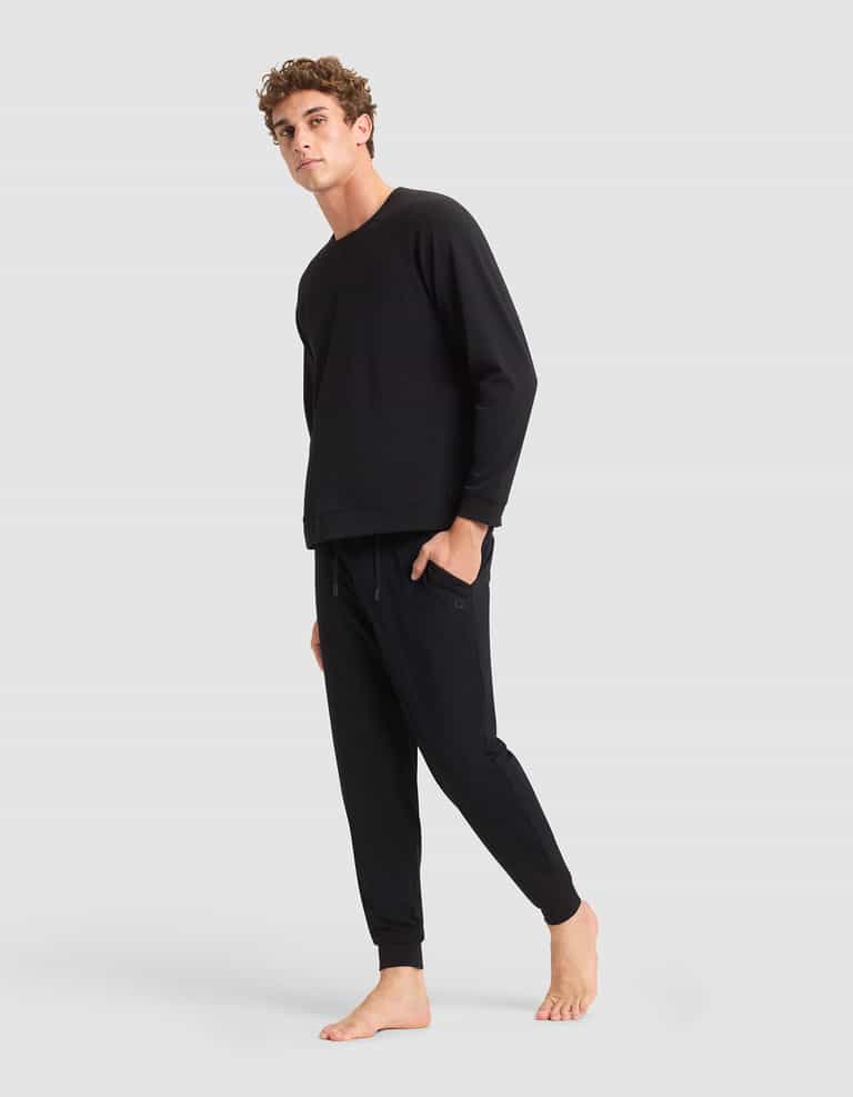 A person stands barefoot against a light gray background, wearing a matching black long-sleeve top and the Men's Brushed Bamboo Jogger Pant by Cozy Earth, looking slightly to the side with one foot raised.