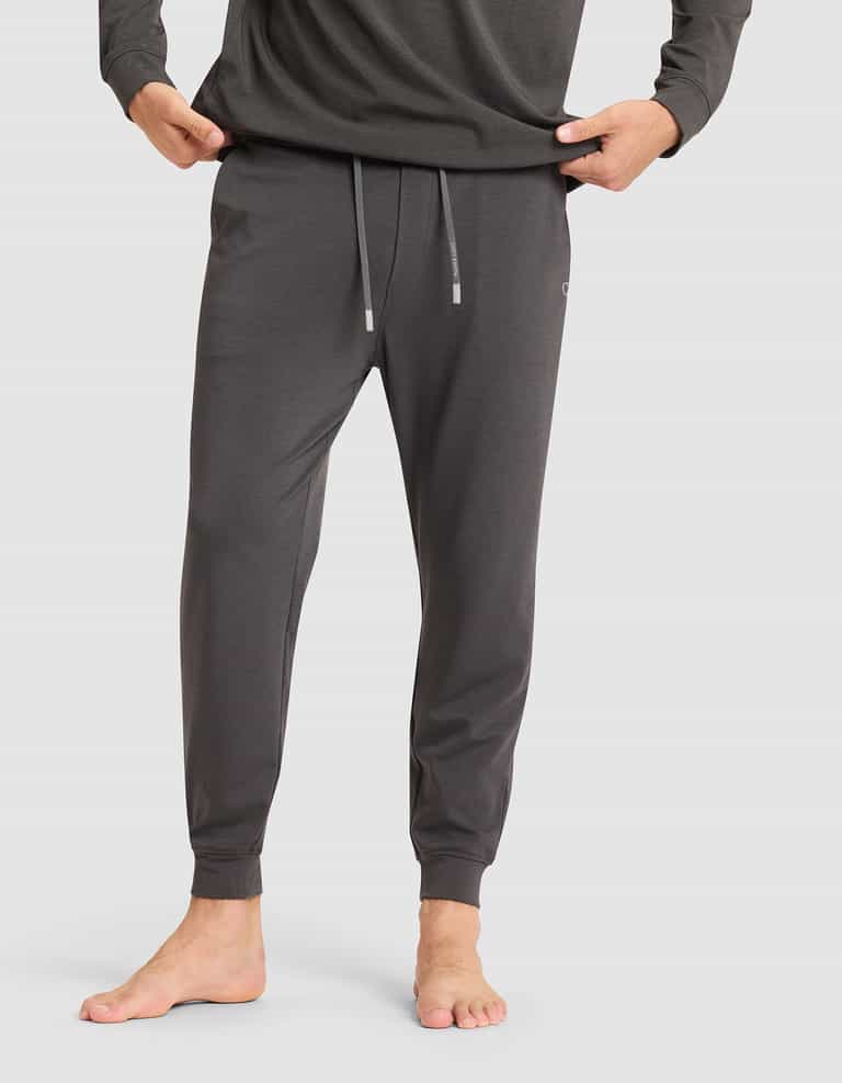 A man stands barefoot against a plain light background, wearing Cozy Earth's Men's Brushed Bamboo Jogger Pant in dark gray with hands in pockets, paired with a matching long-sleeve shirt. |Color:Charcoal