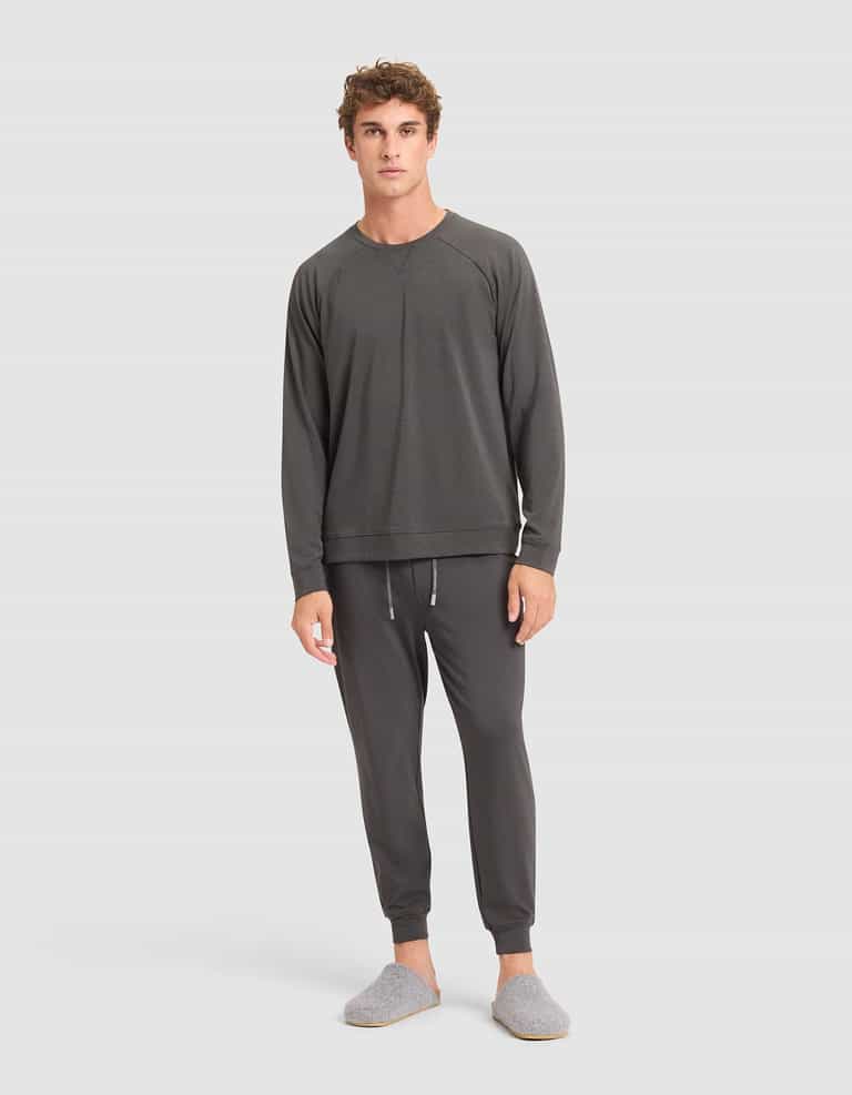 A young man wearing Cozy Earth's Men's Brushed Bamboo Jogger Pant in dark gray, paired with a matching long-sleeve top and gray slippers, stands against a plain white background, looking at the camera. |Color:Charcoal