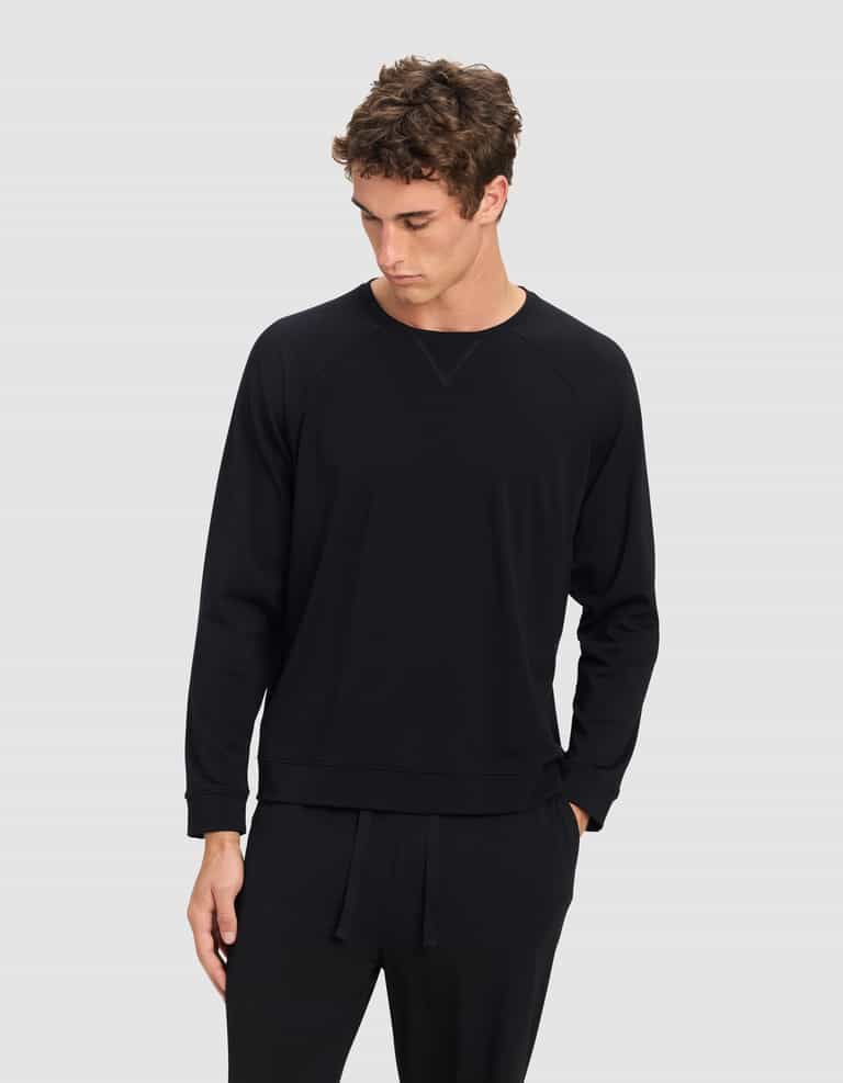 A young man with curly brown hair wears the Cozy Earth Men's Brushed Bamboo Pullover Crew in black, paired with black pants, standing against a plain light grey background and looking down to his left.|Color:Black