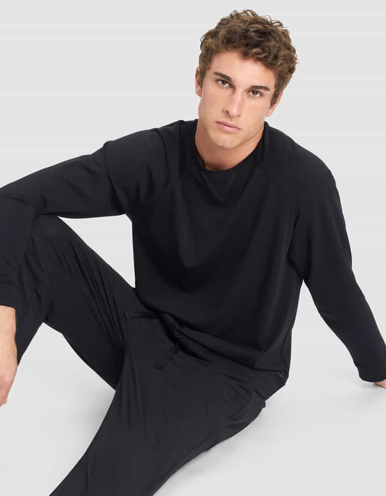 A young man with short curly hair wears the Cozy Earth Men's Brushed Bamboo Pullover Crew and matching pants, sitting on the floor against a light gray background and looking at the camera with a neutral expression.|Color:Black