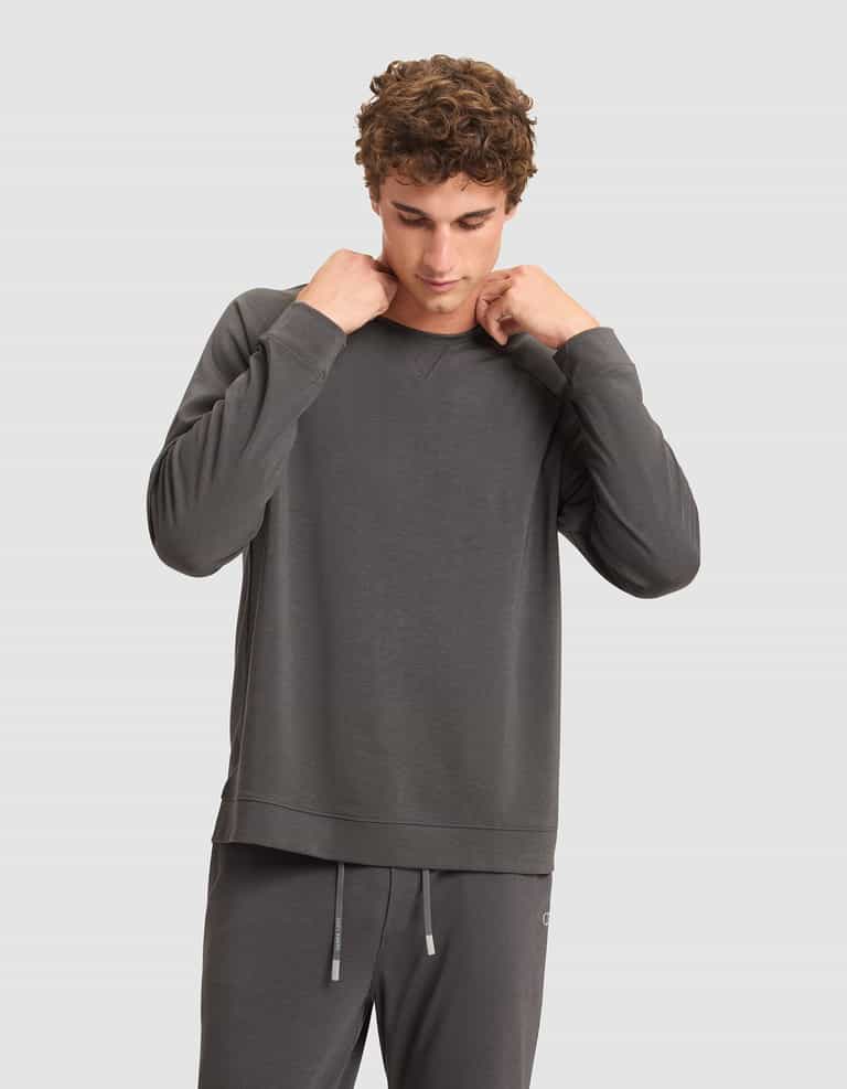 A man with curly hair adjusts the collar of his Cozy Earth Men's Brushed Bamboo Pullover Crew, paired with matching gray pants, while standing against a plain light gray background. |Color:Charcoal