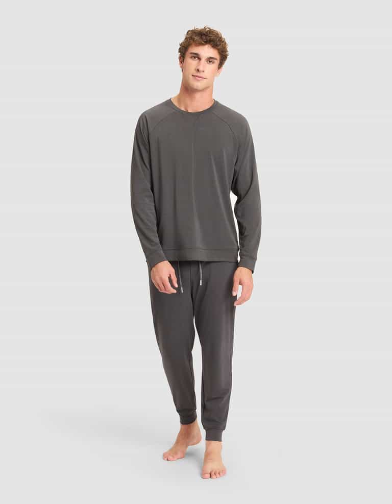 A young man stands barefoot against a plain background, wearing the Cozy Earth Men's Brushed Bamboo Pullover Crew in dark gray lounge set. He appears relaxed, with one hand by his side and the other hanging loosely. |Color:Charcoal
