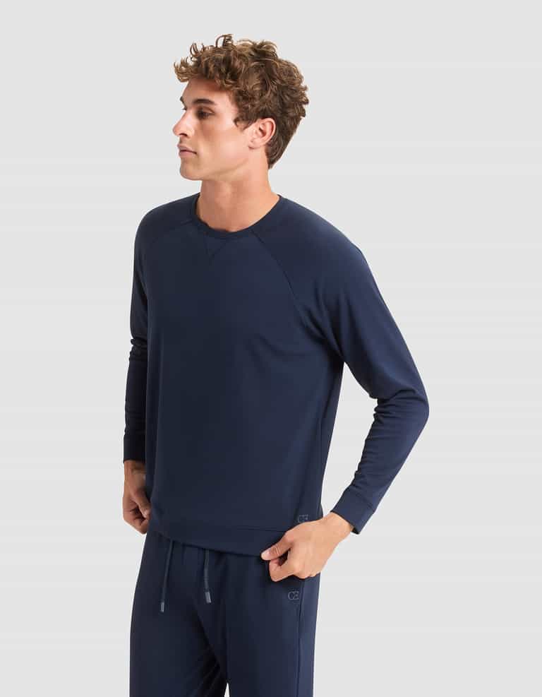 A young man with curly hair, dressed in Cozy Earth's Men's Brushed Bamboo Pullover Crew, stands against a plain light gray background, gazing thoughtfully to the side. |Color:Navy