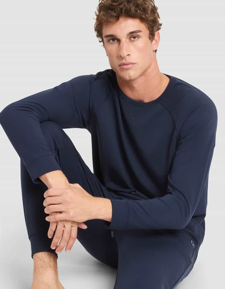 A young man with curly hair wears the Cozy Earth Men's Brushed Bamboo Pullover Crew in navy blue, sitting barefoot on the floor with one arm resting on his knee against a plain light background. |Color:Navy