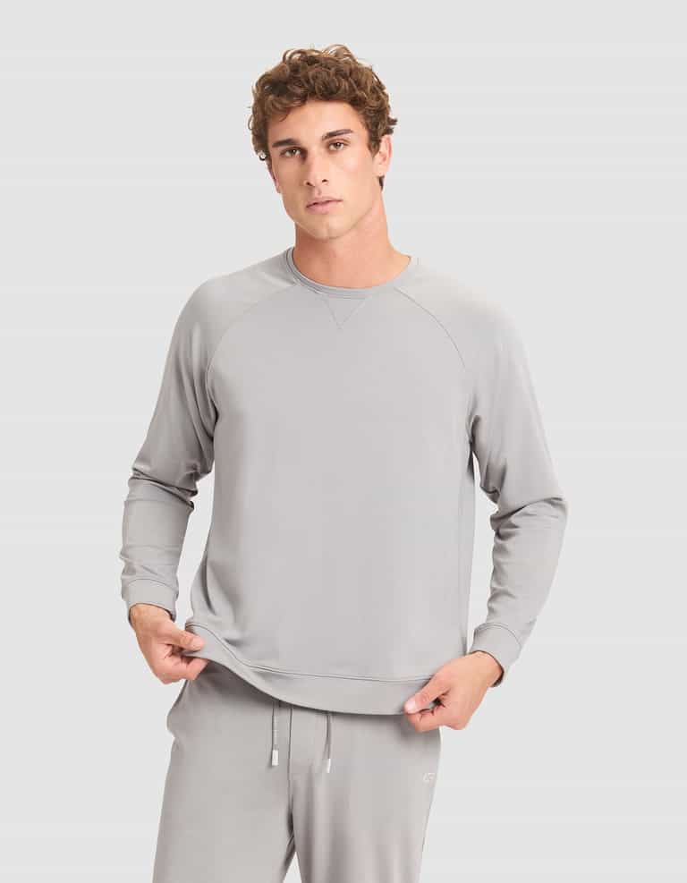 A young man with curly hair wears Cozy Earth's Men's Brushed Bamboo Pullover Crew in light gray, standing against a plain gray background and holding the hem of his sweatshirt. |Color:Stone