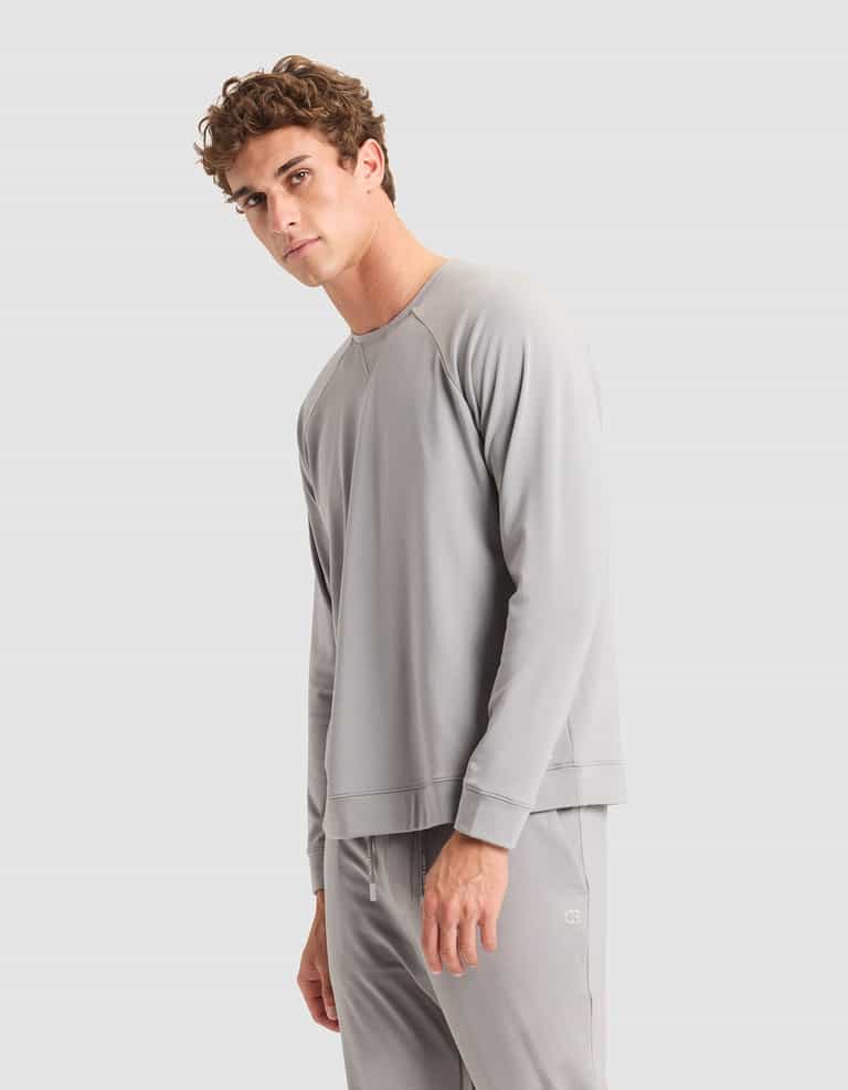A young man with curly brown hair models the Cozy Earth Men's Brushed Bamboo Pullover Crew in light gray, paired with matching pants, standing against a plain light gray background and looking slightly toward the camera with a neutral expression. |Color:Stone