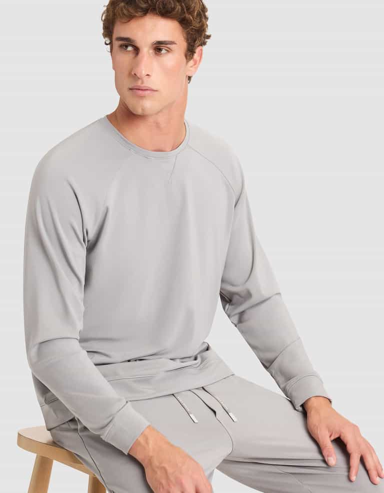 A young man with curly hair, dressed in a light gray Cozy Earth Men's Brushed Bamboo Pullover Crew and matching pants, sits on a wooden stool against a plain light background, gazing slightly to his left. |Color:Stone